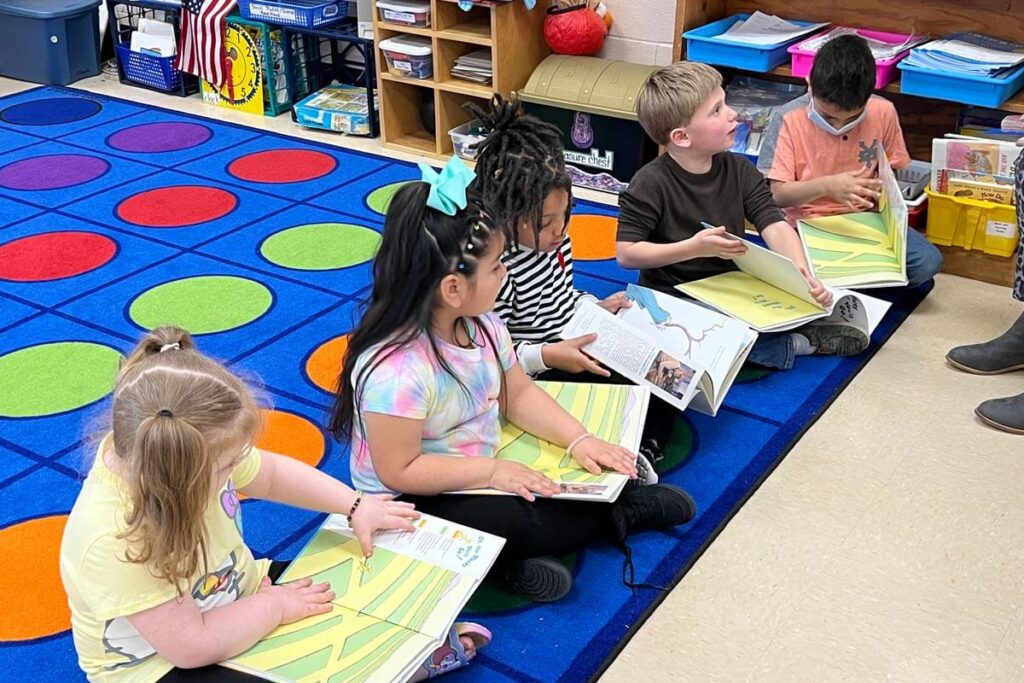 Five children, each with a Dr. Seuss book in their lap, sitting on a brightly colored rug on the floor of a classroom.