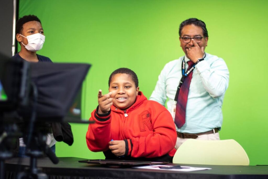 A young student pointing at a video camera while sitting at a desk in front of a green screen as their teacher and another student look on from behind the desk.